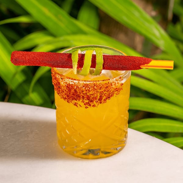 Vibrant yellow cocktail with a chili-lime rim, garnished with lime peels and a red candy straw.