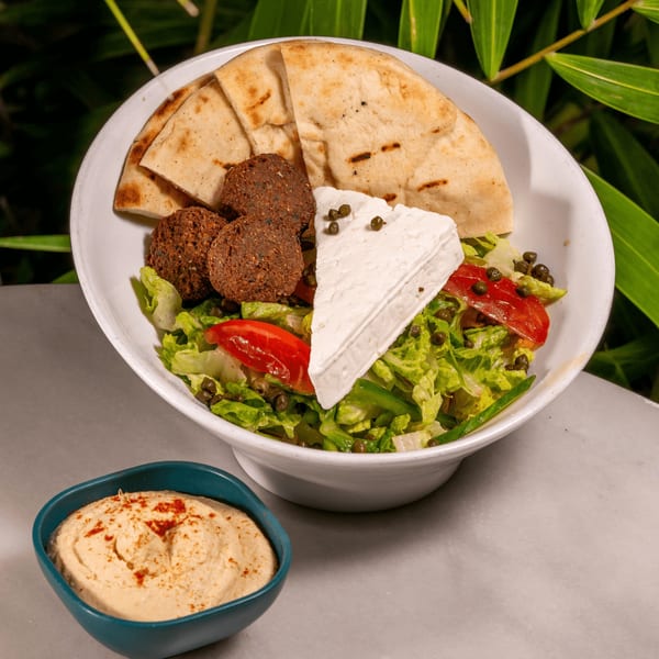 A white bowl of falafel, pita bread, feta cheese, capers, tomatoes, and lettuce, with a side of hummus.