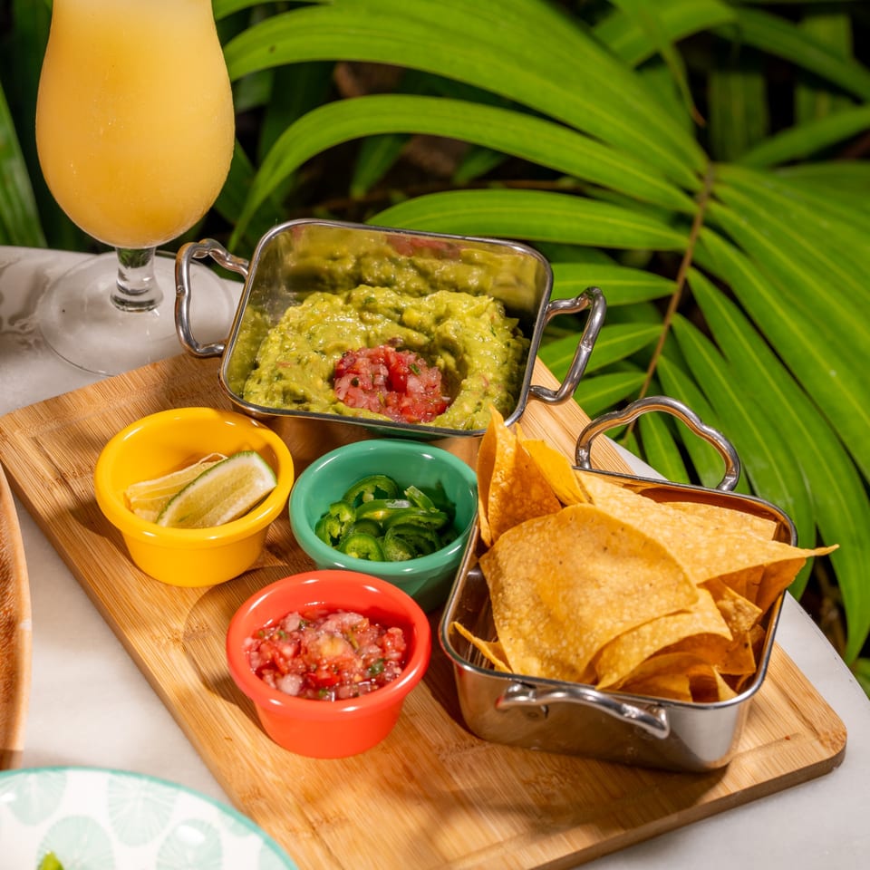 Guacamole, salsa, jalapeños, lime, and tortilla chips on a wooden board with a drink.
