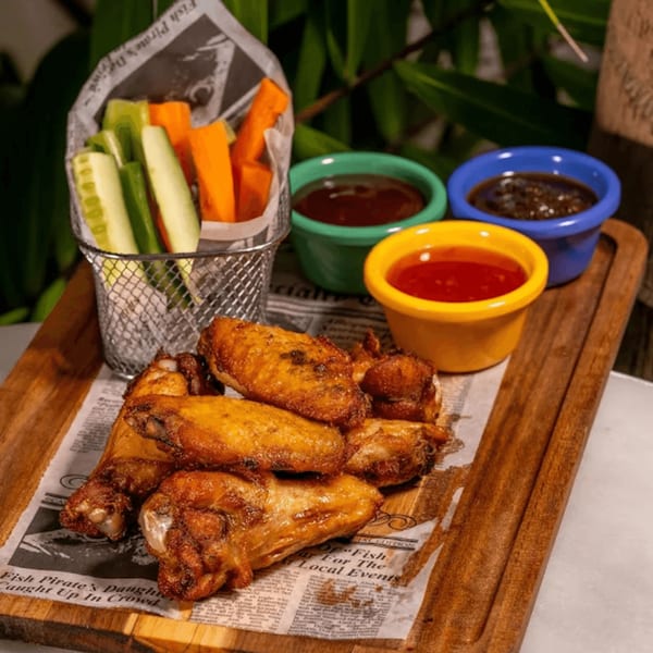 Crispy chicken wings on newspaper, with vegetable sticks and three colorful dipping sauces on a wooden board.