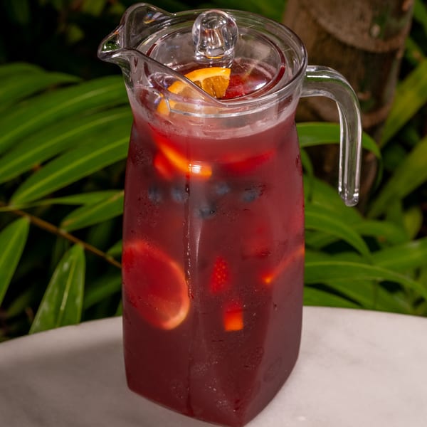 A pitcher of red sangria with fruit slices and berries, set against a lush green background.