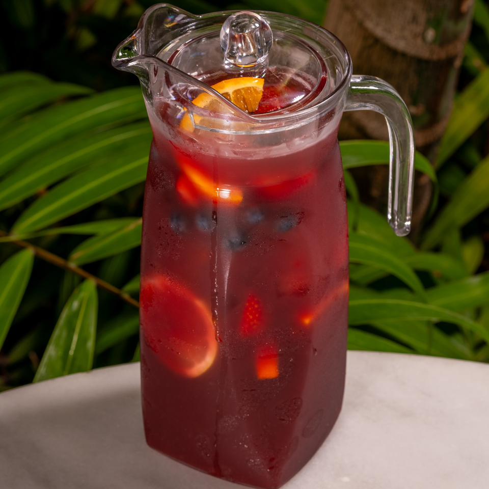 A pitcher of red sangria with fruit slices and berries, set against a lush green background.