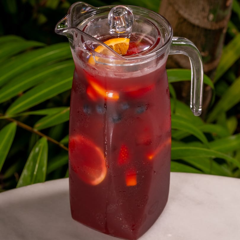 A pitcher of red sangria with fruit slices and berries, set against a lush green background.