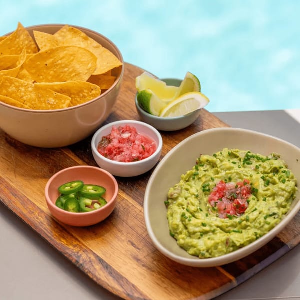 Nachos with guacamole, salsa, sliced jalapeños, and lime wedges on a wooden board by a pool.
