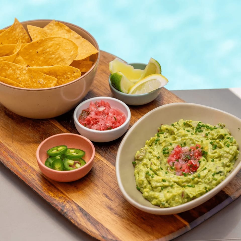 Nachos with guacamole, salsa, sliced jalapeños, and lime wedges on a wooden board by a pool.