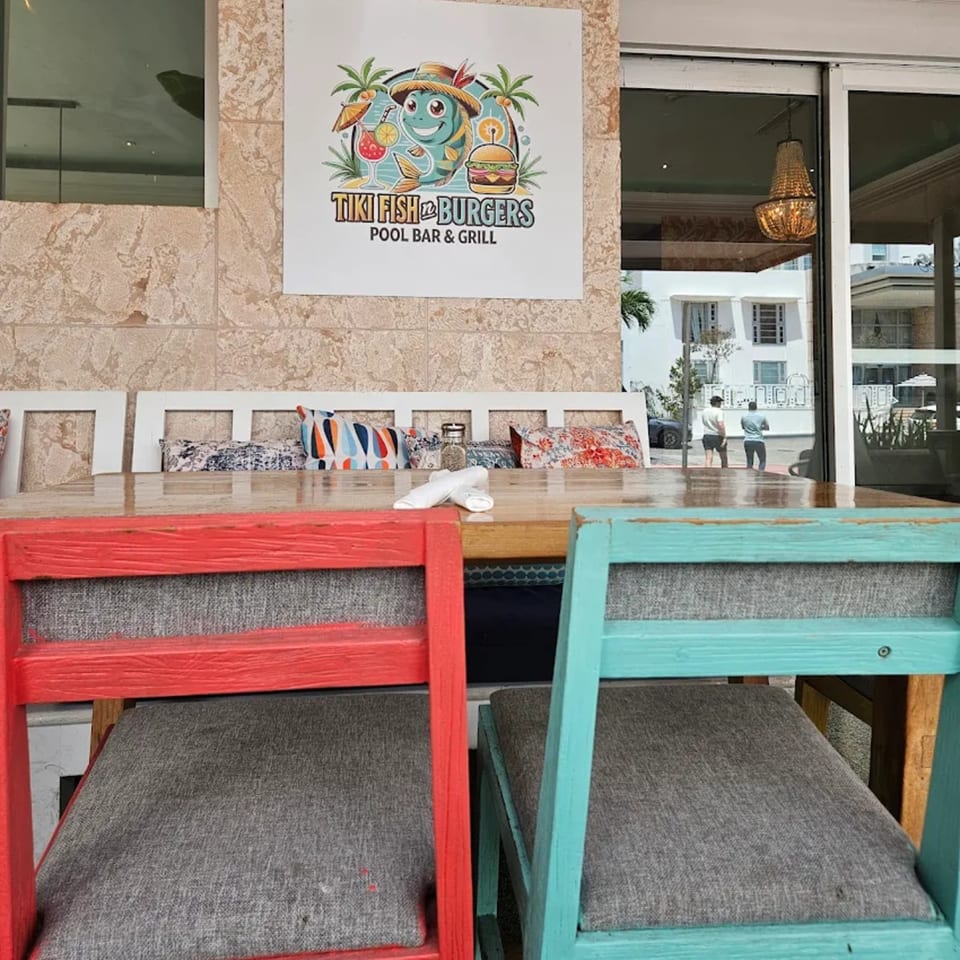 Tiki Fish n Burgers sign above a wooden table with red and teal chairs, looking out to the street.