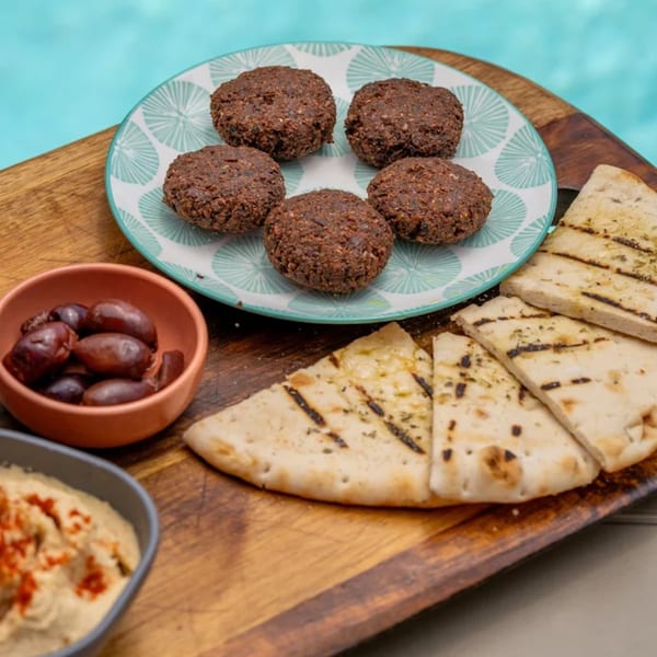 Falafel, grilled pita bread, hummus, and olives on a wooden board by a blue pool.