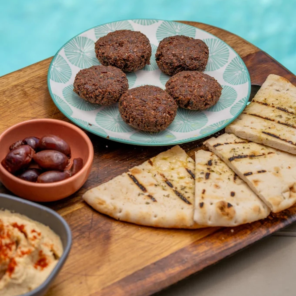 Falafel, grilled pita bread, hummus, and olives on a wooden board by a blue pool.