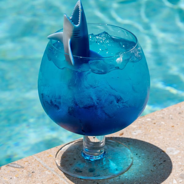 A blue cocktail with ice and a shark toy garnish, sitting on a pool edge.