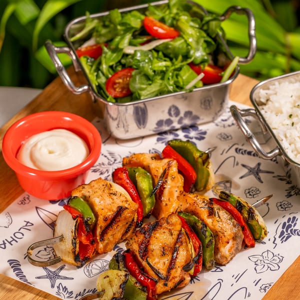 Grilled fish and vegetable skewers with rice, salad, and white sauce on a wooden board.