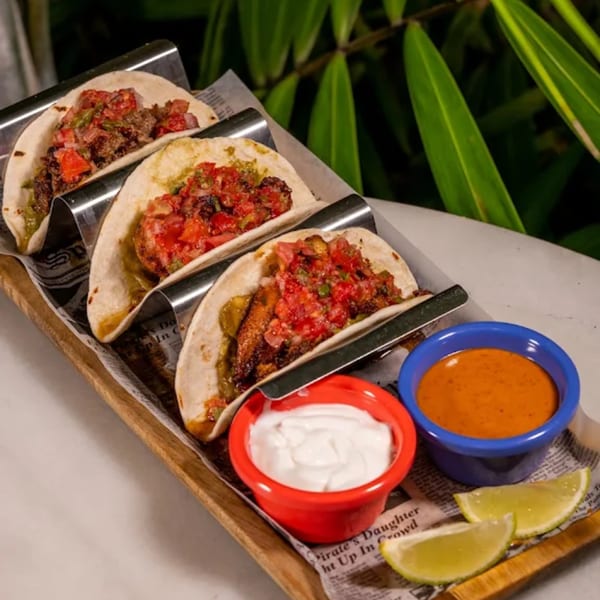Three Tiki Fish n Burgers tacos with salsa, sour cream, and lime wedges on a wooden tray.