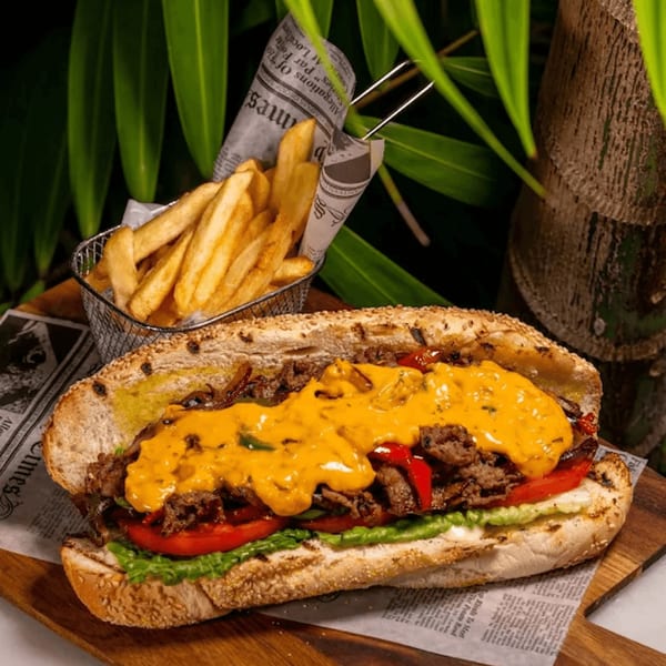 A cheesesteak sandwich with fries in a metal basket, on a wooden board with newspaper.