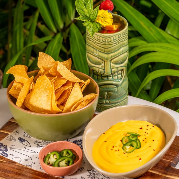 Nachos with cheese dip and jalapeños, alongside a tiki drink, against a lush green background.
